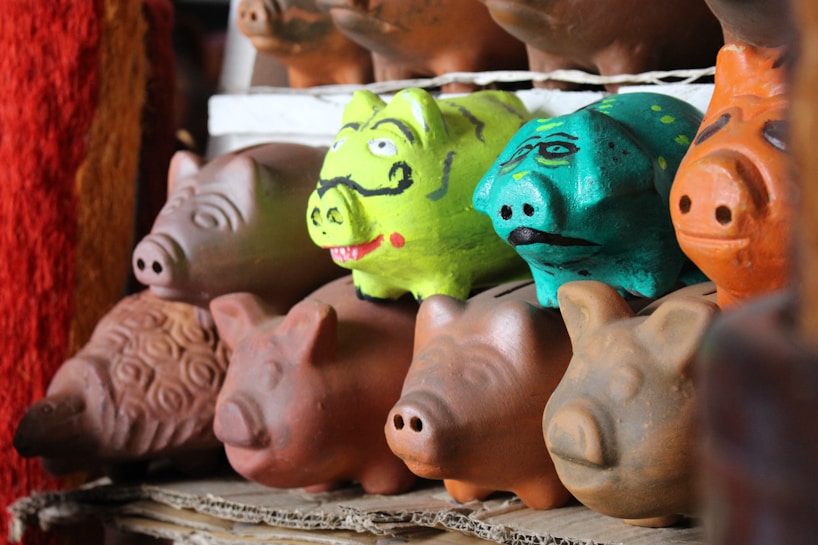 A collection of ceramic piggy banks is arranged on a shelf. The piggy banks display a variety of colors and designs, including plain terracotta colors and vibrant painted patterns. Notably, two piggy banks stand out with bright green and turquoise colors, each with unique facial expressions drawn on them.