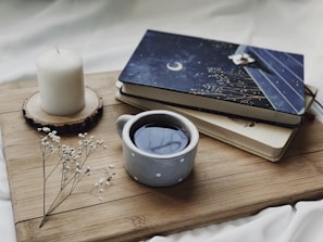 A cozy setting featuring a cup of coffee in a polka dot mug, placed next to two books with a celestial-themed cover on a wooden board. A white candle on a wooden coaster and delicate white flowers add to the tranquil ambiance.