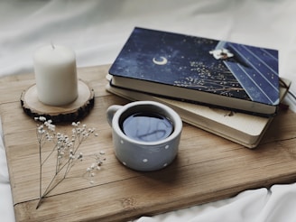 A cozy setting featuring a cup of coffee in a polka dot mug, placed next to two books with a celestial-themed cover on a wooden board. A white candle on a wooden coaster and delicate white flowers add to the tranquil ambiance.