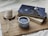 A cozy setting featuring a cup of coffee in a polka dot mug, placed next to two books with a celestial-themed cover on a wooden board. A white candle on a wooden coaster and delicate white flowers add to the tranquil ambiance.