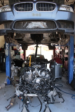 A car is elevated on a hydraulic lift in a garage, revealing its undercarriage. Below, a complex car engine is placed on a stand with numerous cables and components surrounding it. The area is filled with tools and other automotive equipment, creating a busy and industrial atmosphere.