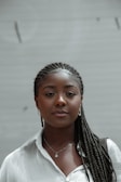 A person with braided hair wearing a white shirt, small hoop earrings, a pendant necklace, and carrying a shoulder bag. The background is a plain, slightly blurred beige surface.