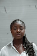 A person with braided hair wearing a white shirt, small hoop earrings, a pendant necklace, and carrying a shoulder bag. The background is a plain, slightly blurred beige surface.