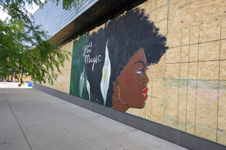 A mural on a boarded-up wall depicts a stylized profile of a woman with dark hair and a prominent earring. The words 'you are Magic' are written on the left side beside a background featuring large white flowers and green leaves.