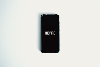 A smartphone with a black screen displaying the word 'INSPIRE' in white text is lying on a plain white surface.