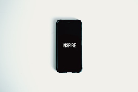 A smartphone with a black screen displaying the word 'INSPIRE' in white text is lying on a plain white surface.