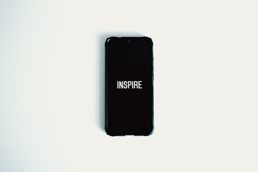 A smartphone with a black screen displaying the word 'INSPIRE' in white text is lying on a plain white surface.