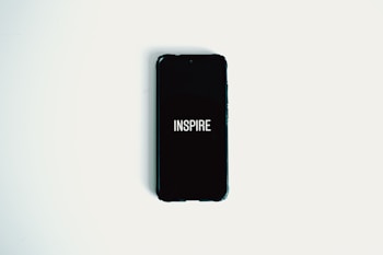 A smartphone with a black screen displaying the word 'INSPIRE' in white text is lying on a plain white surface.