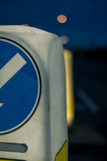 A close-up view of a traffic bollard with a blue, circular directional sign featuring a white arrow. The focus is on the bollard while the background is blurred, showing a dark environment with dim, circular lights possibly from streetlights or vehicles.
