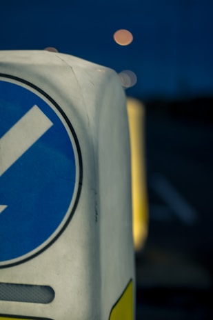 A close-up view of a traffic bollard with a blue, circular directional sign featuring a white arrow. The focus is on the bollard while the background is blurred, showing a dark environment with dim, circular lights possibly from streetlights or vehicles.
