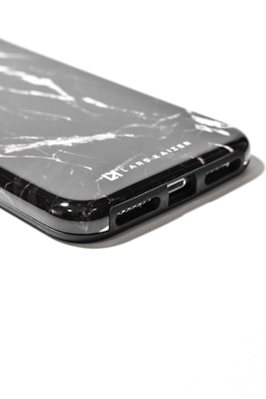 A close-up view of a smartphone case with a black marble pattern and the logo 'LARS KAIZER' engraved on it. The bottom part of the case shows cutouts for speakers and charging port.