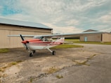A small propeller aircraft with a red and white color scheme is parked on a concrete surface near a hangar. The buildings have beige siding and metal roofs. The background features a partly cloudy sky.