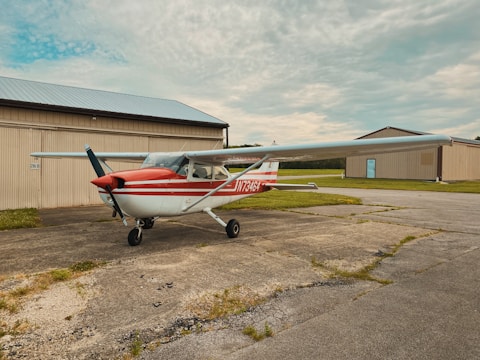 A small propeller aircraft with a red and white color scheme is parked on a concrete surface near a hangar. The buildings have beige siding and metal roofs. The background features a partly cloudy sky.