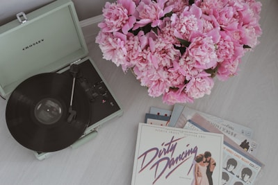 A vintage-style setup featuring a Crosley record player next to a large bouquet of pink peonies. Several vinyl records, including one with 'Dirty Dancing' on the cover, are spread out on the floor. The overall setting suggests a retro and nostalgic vibe.