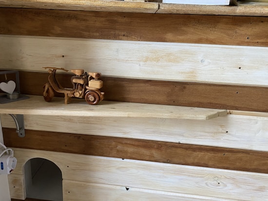 A wooden shelf is adorned with a small, intricately carved wooden motorbike model. The shelf background consists of alternating light and dark wood panels, adding a rustic charm. To the left, there's a heart-shaped metal ornament resting on the shelf.