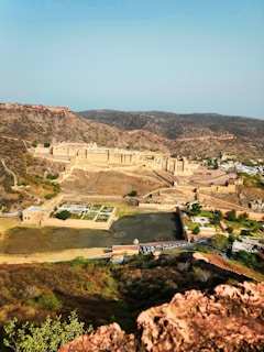 An expansive aerial view of an ancient fort situated on a rugged landscape. The fort, constructed with a distinctive yellow sandstone, is surrounded by arid hills and sparse vegetation. A large courtyard and several architectural structures can be seen within the fort's walls. Below the fort, there is a small water body alongside green patches of garden space.