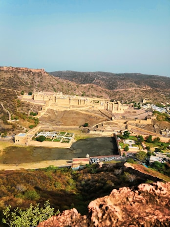 An expansive aerial view of an ancient fort situated on a rugged landscape. The fort, constructed with a distinctive yellow sandstone, is surrounded by arid hills and sparse vegetation. A large courtyard and several architectural structures can be seen within the fort's walls. Below the fort, there is a small water body alongside green patches of garden space.