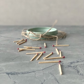 A collection of matches scattered on a gray marble surface. In the background, there is a round, light green ceramic dish wrapped with twine.