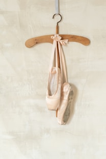A pair of worn ballet pointe shoes with long ribbons are elegantly draped over a wooden hanger. The background features a softly textured light beige or cream-colored wall, adding an understated contrast to the soft pink tones of the ballet shoes.