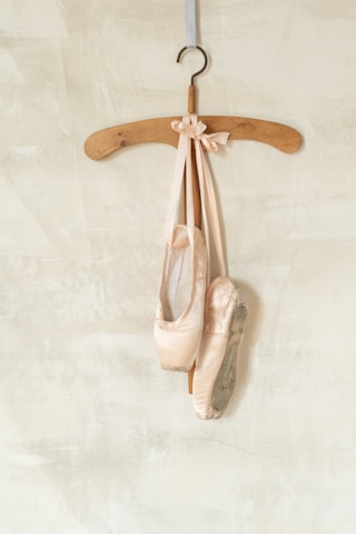 A pair of worn ballet pointe shoes with long ribbons are elegantly draped over a wooden hanger. The background features a softly textured light beige or cream-colored wall, adding an understated contrast to the soft pink tones of the ballet shoes.