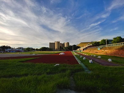 A sports track with red lanes is surrounded by green grass and benches. In the background, a multi-story building and a blue sky with wispy clouds create a serene setting. White bags are placed along the edges of the track.