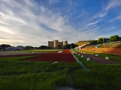 A sports track with red lanes is surrounded by green grass and benches. In the background, a multi-story building and a blue sky with wispy clouds create a serene setting. White bags are placed along the edges of the track.