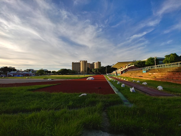 A sports track with red lanes is surrounded by green grass and benches. In the background, a multi-story building and a blue sky with wispy clouds create a serene setting. White bags are placed along the edges of the track.