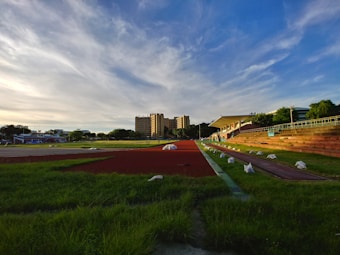 A sports track with red lanes is surrounded by green grass and benches. In the background, a multi-story building and a blue sky with wispy clouds create a serene setting. White bags are placed along the edges of the track.