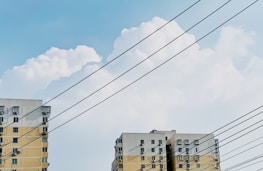 Two residential buildings with multiple windows are situated against a backdrop of white, fluffy clouds in a blue sky. Several parallel power lines cut across the view.