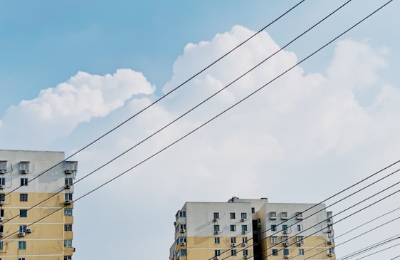 Two residential buildings with multiple windows are situated against a backdrop of white, fluffy clouds in a blue sky. Several parallel power lines cut across the view.