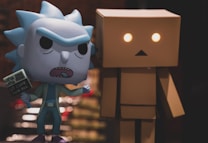 Two toy figures are displayed, one resembling a humanoid character with blue spiky hair and an exaggerated facial expression, holding a small placard. The other is a boxy figure with glowing eyes, featuring a simple, geometric design. The background is blurred, emphasizing the two figures in the foreground.