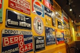 A wall is covered with a variety of vintage metal advertising and street signs. These signs include a mix of colors and fonts, featuring brands like Pepsi-Cola and Ricard, as well as street names and business advertisements. The setting has a warm ambiance with yellow lighting reflecting off the signs.