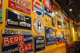 A wall is covered with a variety of vintage metal advertising and street signs. These signs include a mix of colors and fonts, featuring brands like Pepsi-Cola and Ricard, as well as street names and business advertisements. The setting has a warm ambiance with yellow lighting reflecting off the signs.