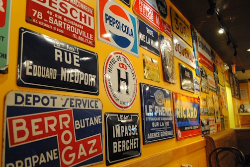 A wall is covered with a variety of vintage metal advertising and street signs. These signs include a mix of colors and fonts, featuring brands like Pepsi-Cola and Ricard, as well as street names and business advertisements. The setting has a warm ambiance with yellow lighting reflecting off the signs.
