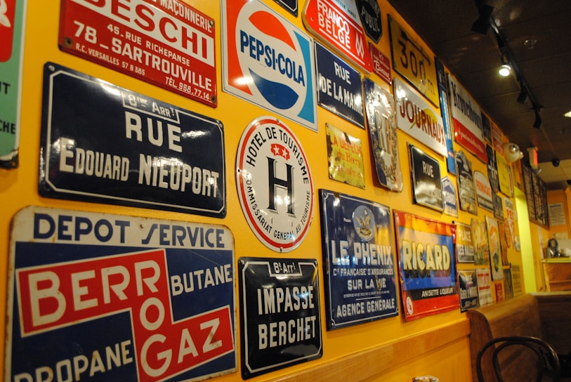 A wall is covered with a variety of vintage metal advertising and street signs. These signs include a mix of colors and fonts, featuring brands like Pepsi-Cola and Ricard, as well as street names and business advertisements. The setting has a warm ambiance with yellow lighting reflecting off the signs.