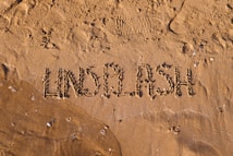 The image features the word 'UNSPLASH' written in the wet sand on a beach. The letters are formed by slight indentations in the sand, and a few small pebbles and shells are scattered around. Footprints are visible in the background, suggesting recent human activity. The sunlight casts soft shadows, enhancing the texture of the sand.