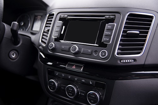 The interior of a car's dashboard featuring a central console with a multimedia display and various control buttons including navigation and setup options. There are several knobs and buttons for adjusting audio and climate settings. The dashboard is sleek, with air vents and a modern, polished finish.