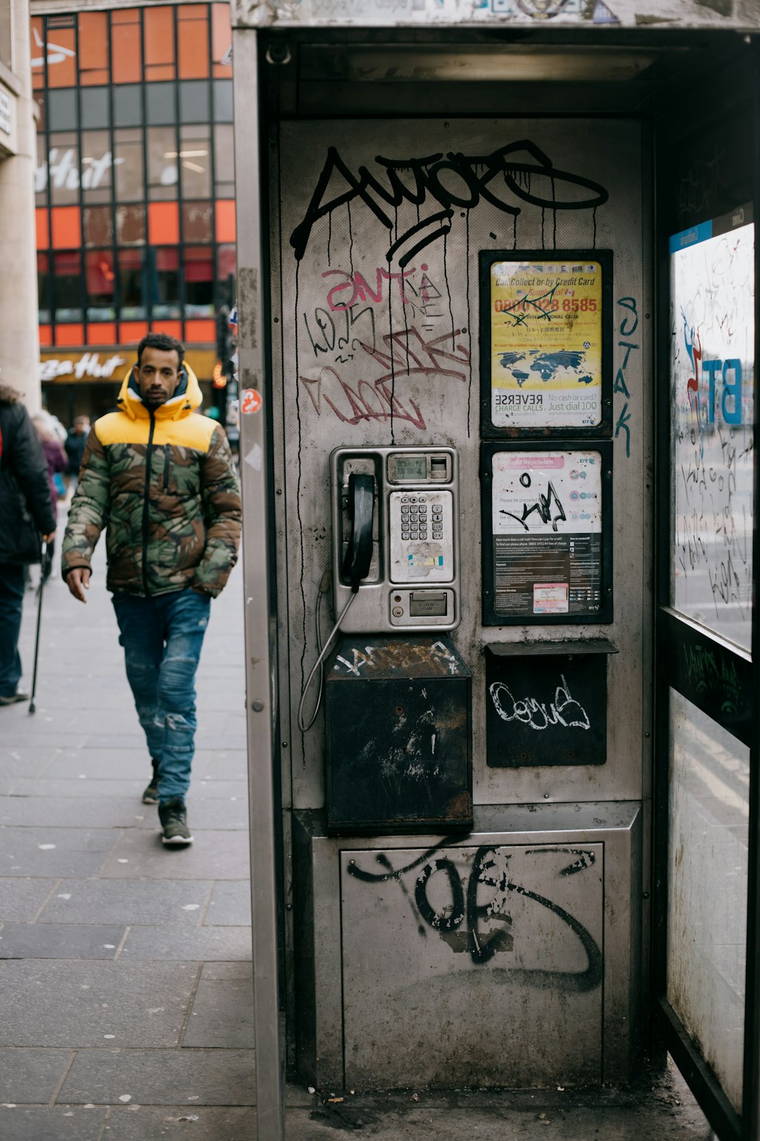 man in yellow jacket and blue denim jeans standing beside black telephone booth