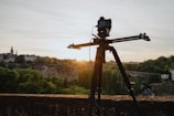 A camera mounted on a track slider stands on a tripod overlooking a scenic landscape. The background features a historic town with old buildings and lush green trees under a clear sky at sunset.