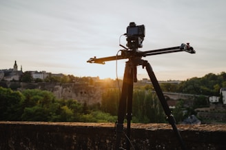 A camera mounted on a track slider stands on a tripod overlooking a scenic landscape. The background features a historic town with old buildings and lush green trees under a clear sky at sunset.