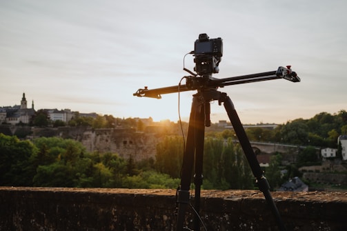 A camera mounted on a track slider stands on a tripod overlooking a scenic landscape. The background features a historic town with old buildings and lush green trees under a clear sky at sunset.