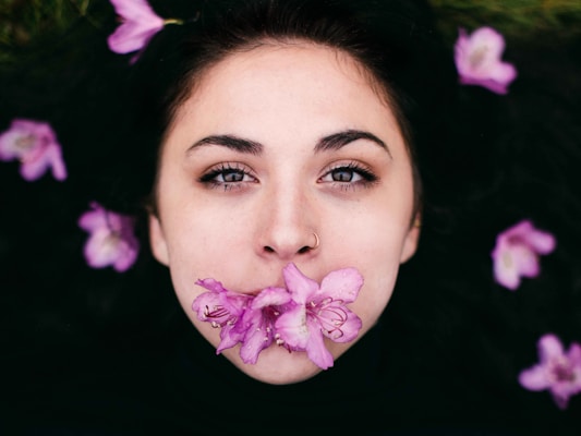 A close-up of a person's face with light skin, piercing eyes, and a nose ring. Pink flowers cover the mouth and scatter around the head on a dark background, creating a striking composition.