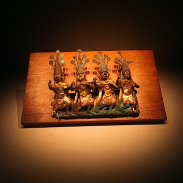 A carved wooden plaque featuring four ancient-looking bronze figures with elaborate headdresses. The figures appear to be in a dance or ritualistic pose and are set against a wooden background.