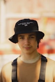 A person wearing a black bucket hat with text 'THE HUNDREDS SINCE 1980' and a beige shirt with black suspenders. The background is blurred, featuring warm tones.
