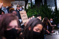 A group of people are gathered, some sitting and some standing, with several individuals holding protest signs. One prominent sign reads 'My skin color is not a crime threat.' Many are wearing masks and appear to be attentively listening or engaged in the demonstration. There is a sense of unity and focus in the group.