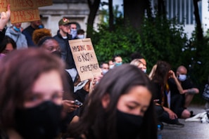 A group of people are gathered, some sitting and some standing, with several individuals holding protest signs. One prominent sign reads 'My skin color is not a crime threat.' Many are wearing masks and appear to be attentively listening or engaged in the demonstration. There is a sense of unity and focus in the group.