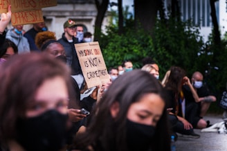 A group of people are gathered, some sitting and some standing, with several individuals holding protest signs. One prominent sign reads 'My skin color is not a crime threat.' Many are wearing masks and appear to be attentively listening or engaged in the demonstration. There is a sense of unity and focus in the group.