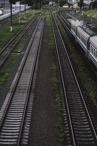 A railway scene featuring multiple train tracks running parallel. On the right, part of a train is visible, moving along one of the tracks. The tracks are bordered by green grass and small patches of vegetation. Overhead cables and poles for the train electrification system are present. In the distance, buildings and a station platform can be seen.