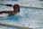 A swimmer wearing a red swim cap and goggles is performing a swimming stroke in a pool. Water splashes around them as they move forward, and lane dividers are visible in the pool.