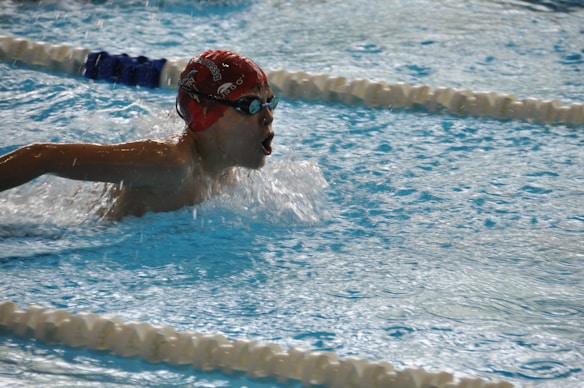 A swimmer wearing a red swim cap and goggles is performing a swimming stroke in a pool. Water splashes around them as they move forward, and lane dividers are visible in the pool.